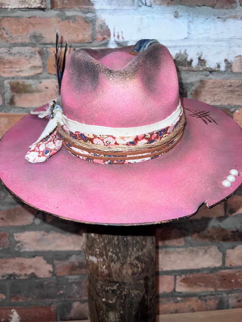May include: A pink felt cowboy hat with a floral patterned band, a feather, and a denim ribbon. The hat has a distressed look with white paint splatters.