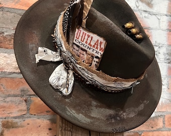 May include: A distressed brown cowboy hat with decorative feathers, a barbed wire band, and a small sign that reads "Outlaw Country." The hat is adorned with a white patterned scarf and brass-colored accents.