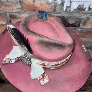 May include: A pink cowboy hat with a distressed finish, featuring a denim band, a white feather, and a decorative fabric bow with a rhinestone embellishment.
