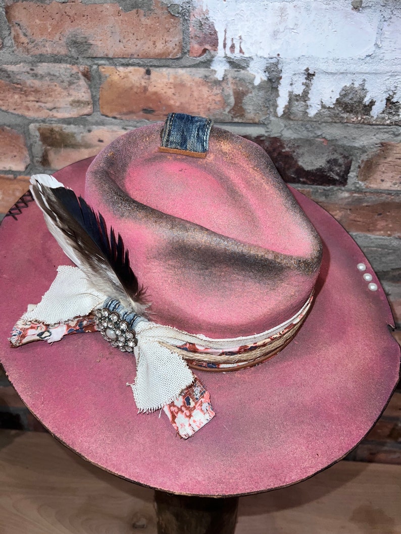 May include: A pink cowboy hat with a distressed finish, featuring a denim band, a white feather, and a decorative fabric bow with a rhinestone embellishment.