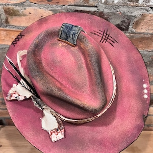 May include: A pink felt fedora hat with a distressed look. The hat has a denim patch on the top, a white ribbon band, and a feather accent.