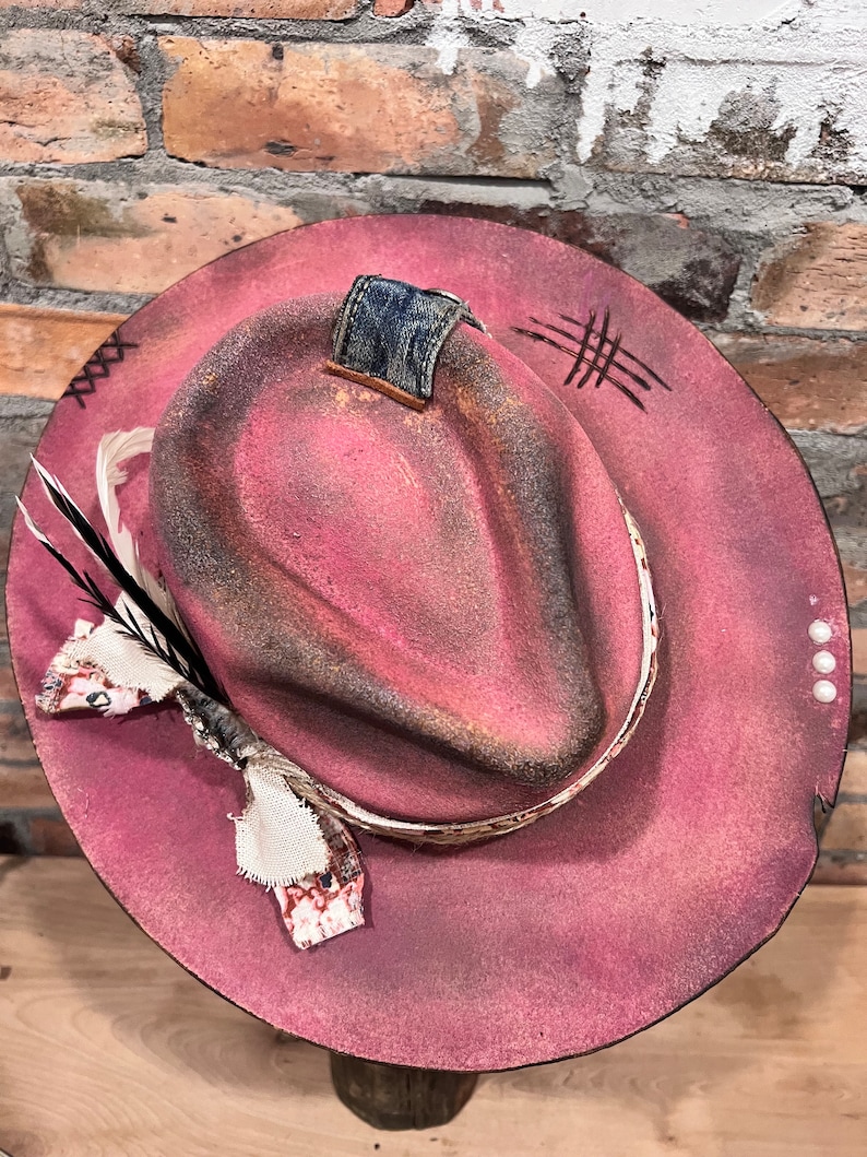 May include: A pink felt fedora hat with a distressed look. The hat has a denim patch on the top, a white ribbon band, and a feather accent.