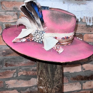 May include: A pink cowboy hat with a white and floral patterned band, a white feather, and a rhinestone pin. The hat is sitting on a wooden post.