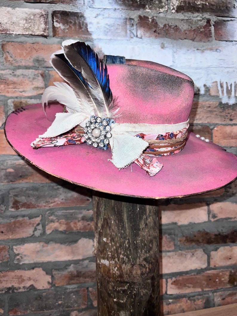 May include: A pink cowboy hat with a white and floral patterned band, a white feather, and a rhinestone pin. The hat is sitting on a wooden post.