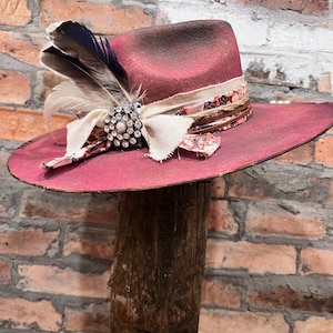 May include: A pink felt cowboy hat with a brown leather band and a white and floral ribbon. The hat has a feather and a rhinestone brooch.