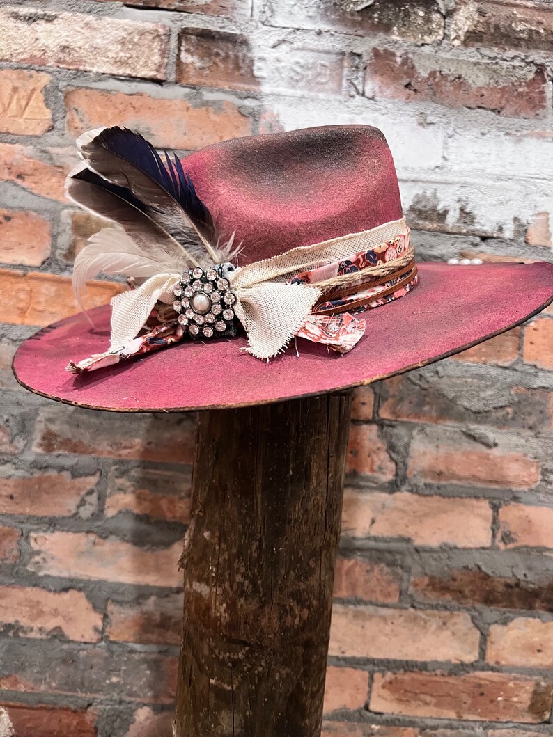 May include: A pink felt cowboy hat with a brown leather band and a white and floral ribbon. The hat has a feather and a rhinestone brooch.