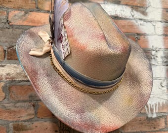 May include: A beige cowboy hat with a gold chain, blue and beige ribbon, and feather accents. The hat has a distressed, painted look with red, blue, and purple hues. A tag with text is attached to the hat.