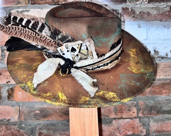 May include: A brown felt cowboy hat with a distressed look. The hat is decorated with a feather, a playing card, and a chain. The hat is sitting on a wooden stand.