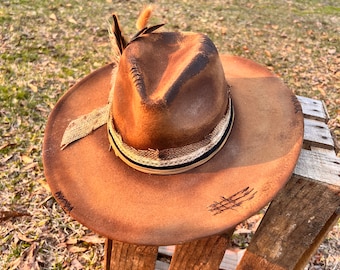 May include: Brown fedora-style hat with a distressed look. The hat features a decorative band with a burlap ribbon and a black and white stripe. A feather is attached to the side. The hat is sitting on a wooden pallet.