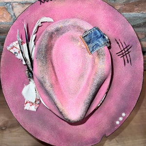 May include: A pink cowboy hat with a denim patch, a feather, and white beads around the brim.