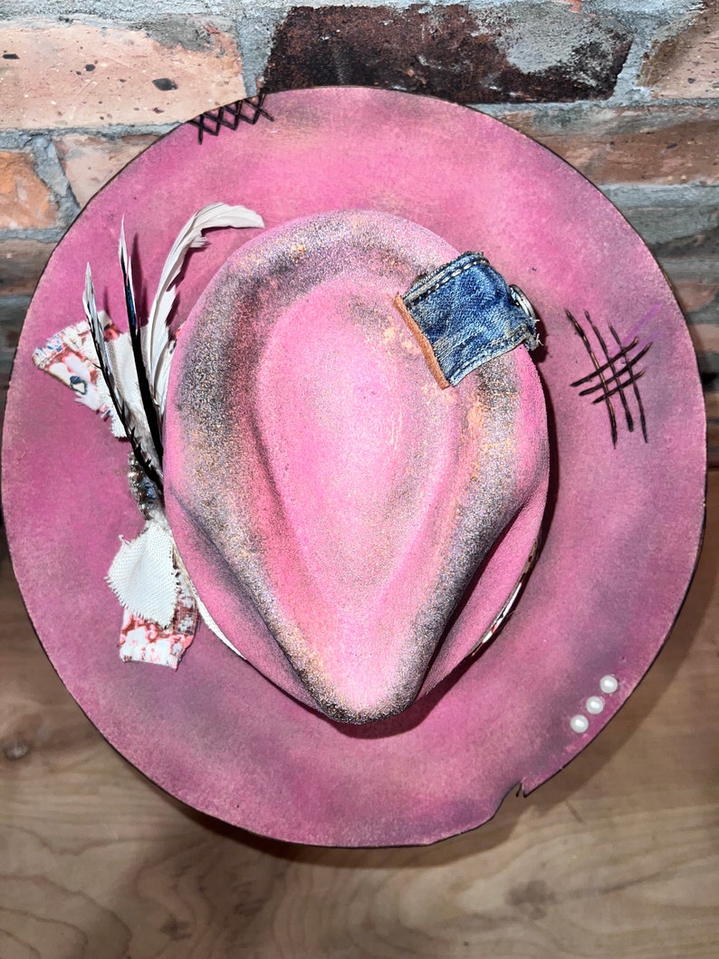 May include: A pink cowboy hat with a denim patch, a feather, and white beads around the brim.