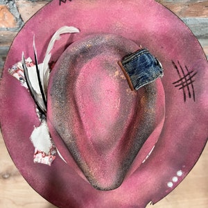 May include: A pink felt fedora hat with a distressed look. The hat has a denim band and a feather accent.