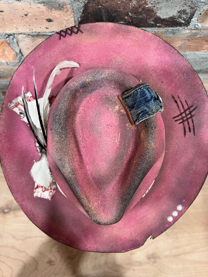 May include: A pink felt fedora hat with a distressed look. The hat has a denim band and a feather accent.