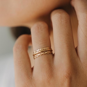 May include: Close-up of three gold rings stacked on a finger. The rings feature different designs: a dotted band, a wavy band, and a simple band. The rings are a warm gold color and appear delicate.