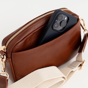 May include: A brown leather crossbody bag with a cream canvas strap and a gold zipper. A black smartphone is inside the bag with the camera lens visible.
