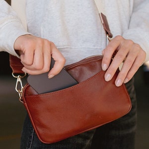 May include: A brown leather crossbody bag with a white strap. A person is holding a black phone in the bag.