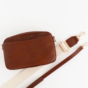 May include: A brown leather crossbody bag with a white and brown adjustable strap. The bag has a flap closure and a small pocket on the front.