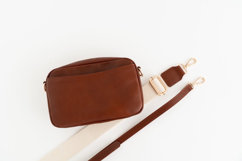 May include: A brown leather crossbody bag with a white and brown adjustable strap. The bag has a flap closure and a small pocket on the front.