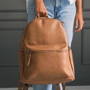 May include: A tan backpack with a front zipped pocket and adjustable shoulder straps. The backpack is made of a smooth material and has a top handle. The hardware is bronze-coloured.