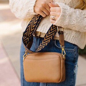 May include: A brown leather crossbody bag with a gold zipper and a navy blue and gold chevron patterned strap.