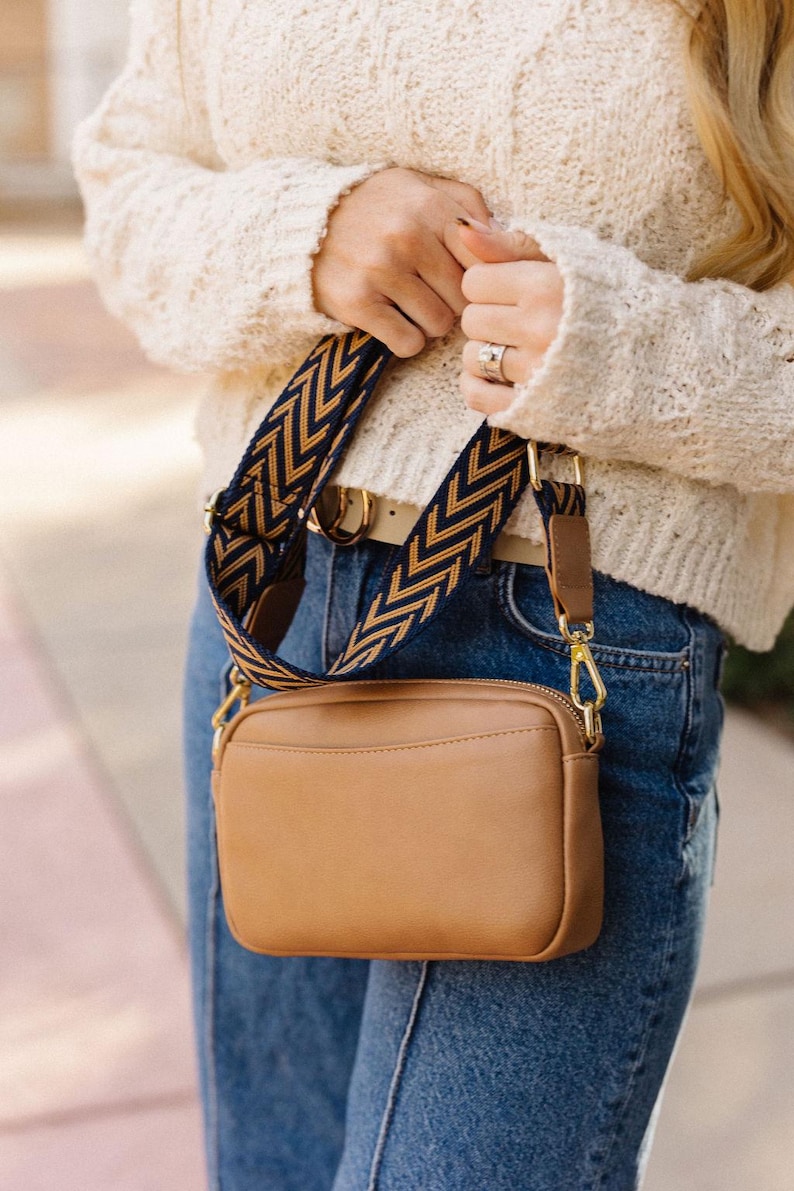 May include: A brown leather crossbody bag with a gold zipper and a navy blue and gold chevron patterned strap.