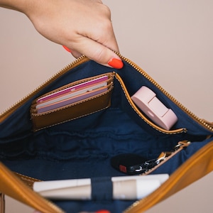 May include: A tan leather clutch with a navy blue interior, open to reveal a brown card holder, a pink item, keys, and a tube of lip balm. The clutch is held open by a hand with orange nail polish.