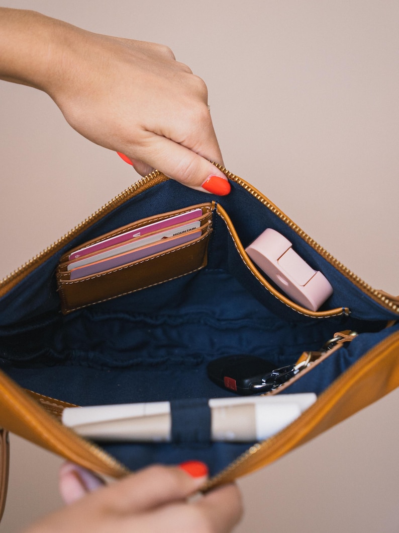 May include: A tan leather clutch with a navy blue interior, open to reveal a brown card holder, a pink item, keys, and a tube of lip balm. The clutch is held open by a hand with orange nail polish.