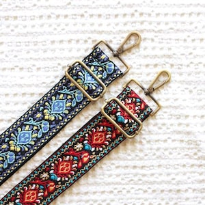 May include: Two decorative bag straps with brass-colored hardware. One strap is black with blue, yellow, and green floral embroidery. The other is black with red, turquoise, and white embroidered patterns. Both straps have adjustable buckles and swivel clasps.
