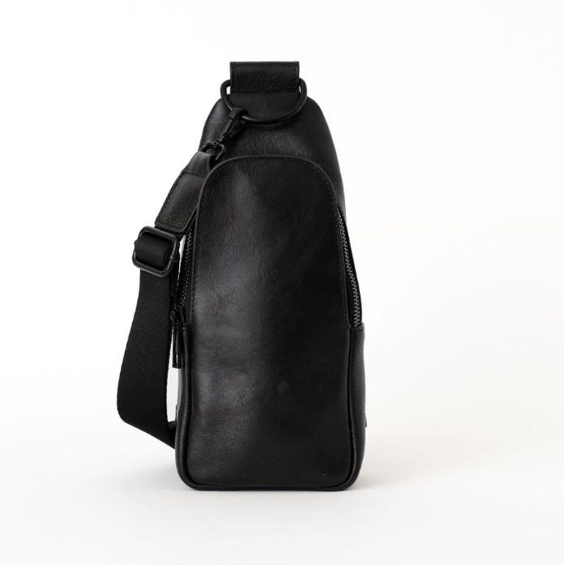 May include: A black sling bag with a single adjustable shoulder strap. The bag has a zippered front pocket and a top handle. The bag is made of a smooth, leather-like material and is designed to be worn across the body.
