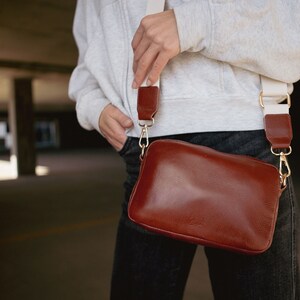 May include: A brown leather crossbody bag with a white strap. The bag has a flap closure and a gold buckle.