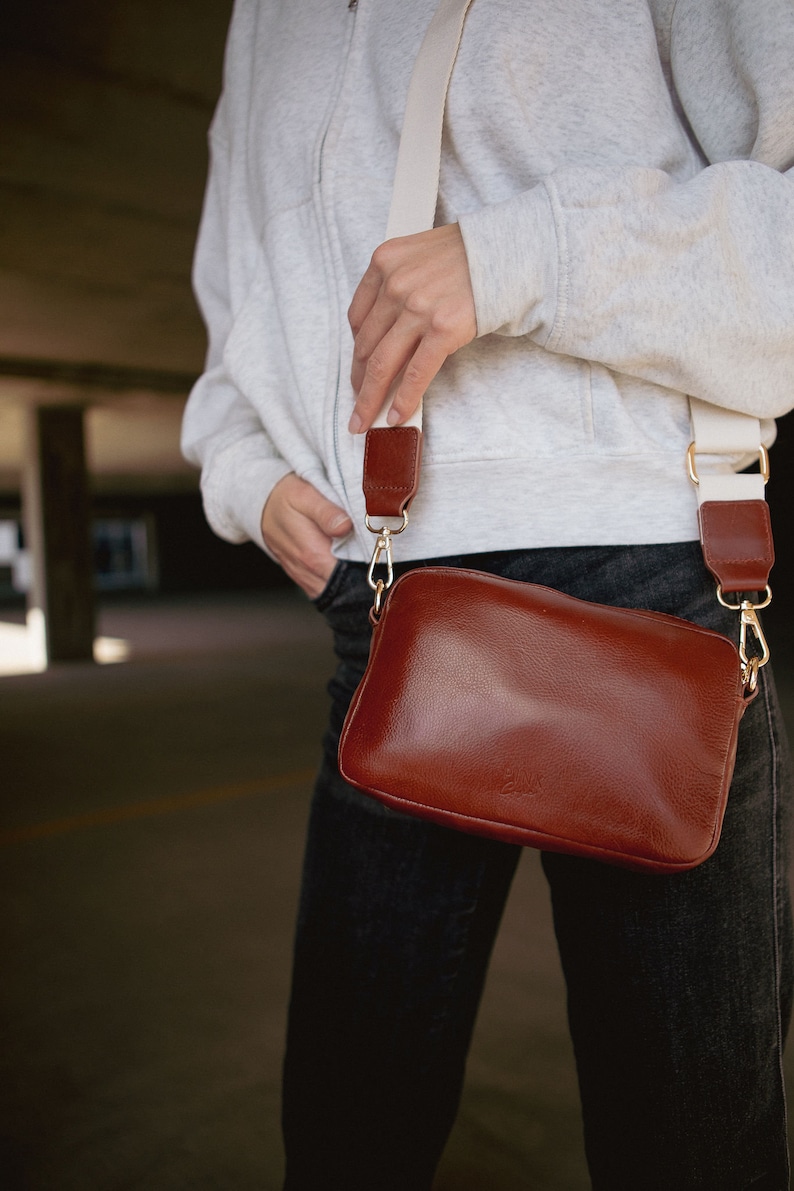 May include: A brown leather crossbody bag with a white strap. The bag has a flap closure and a gold buckle.