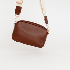 May include: A brown leather crossbody bag with a white and brown strap. The bag has a rectangular shape and a gold clasp.