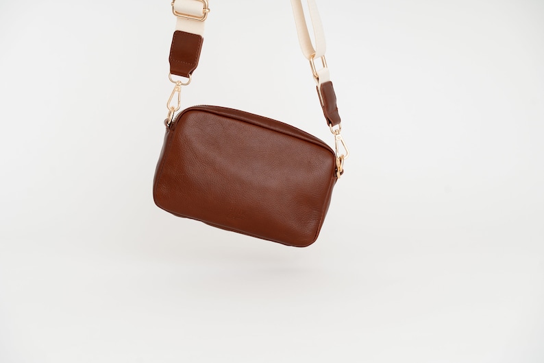 May include: A brown leather crossbody bag with a white and brown strap. The bag has a rectangular shape and a gold clasp.