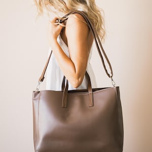 May include: A brown tote bag with a shoulder strap and two top handles. The bag is rectangular and appears to be made of leather or a leather-like material. The bag is being carried over the shoulder.
