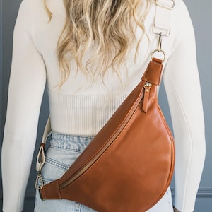 May include: A brown leather fanny pack with a gold zipper and white strap. The bag is worn across the body. The strap is adjustable and connects to the bag with gold-colored hardware. The bag is made of smooth leather.