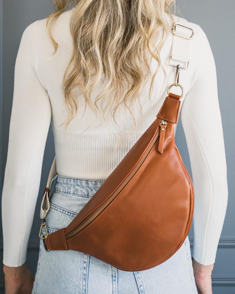 May include: A brown leather fanny pack with a gold zipper and white strap. The bag is worn across the body. The strap is adjustable and connects to the bag with gold-colored hardware. The bag is made of smooth leather.