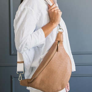 May include: A tan crossbody bag with a textured, grid-like pattern. The bag has a long, adjustable strap with gold-colored hardware and a white section. The bag is worn over the shoulder.