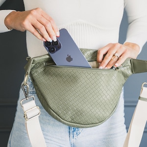 May include: Olive green woven fanny pack with a cream-colored adjustable strap. The bag has a zippered main compartment and a silver-colored clasp. A blue smartphone is placed inside the bag.