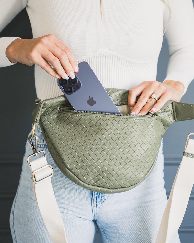 May include: Olive green woven fanny pack with a cream-colored adjustable strap. The bag has a zippered main compartment and a silver-colored clasp. A blue smartphone is placed inside the bag.
