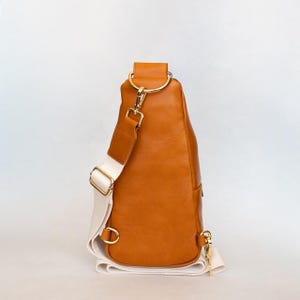 May include: A brown leather sling bag with a white strap and gold-colored hardware. The bag has a rounded shape and a single strap that goes over the shoulder. The bag is approximately 12 inches tall.
