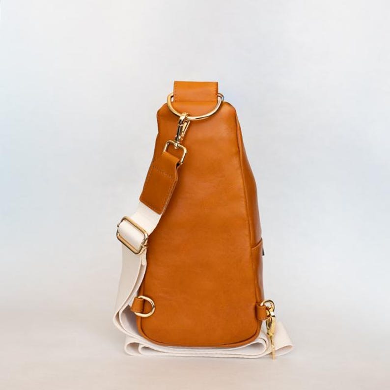 May include: A brown leather sling bag with a white strap and gold-colored hardware. The bag has a rounded shape and a single strap that goes over the shoulder. The bag is approximately 12 inches tall.
