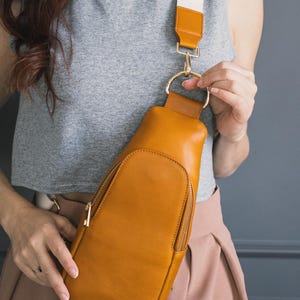 May include: Mustard-colored leather sling bag with a gold zipper and a cream and brown adjustable strap. The bag has a rounded shape and a gold metal ring attachment.