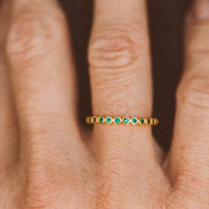 May include: A gold ring with a series of small, round, emerald-green gemstones set in a row. The ring is on a finger, and the background is a blurred, warm tone.