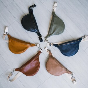 May include: A collection of six crescent-shaped fanny packs in various colors, including black, olive green, tan, brown, and navy blue. Each bag features a zipper closure, adjustable strap, and silver-tone hardware. The bags are arranged in a circular pattern.