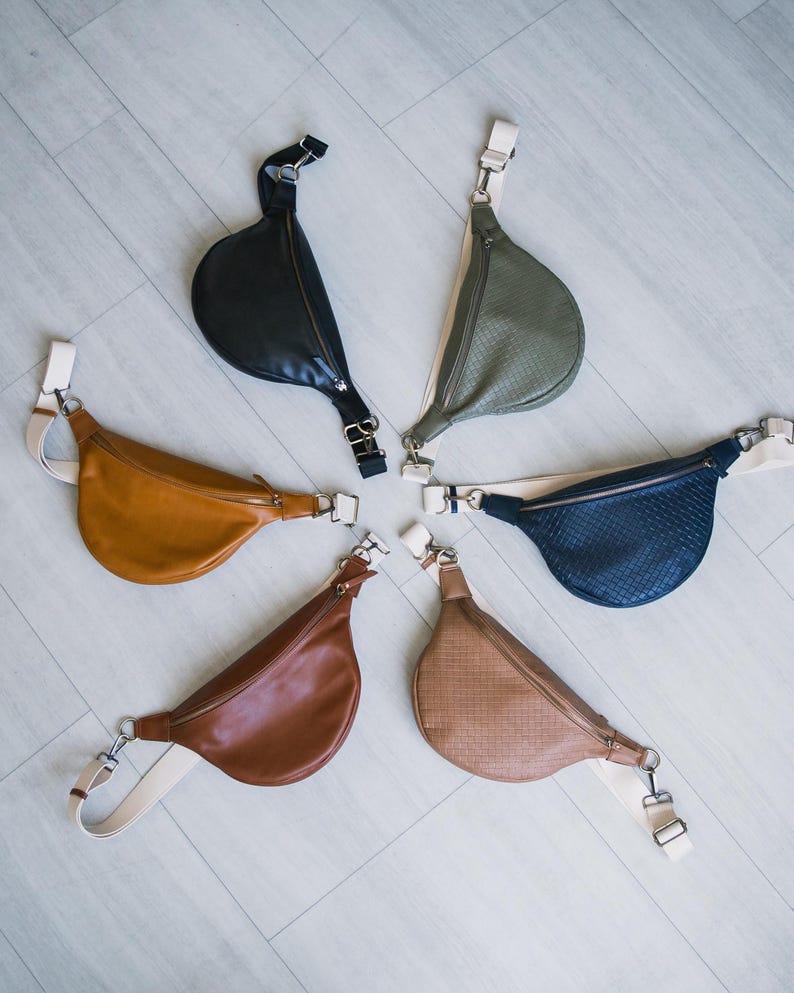 May include: A collection of six crescent-shaped fanny packs in various colors, including black, olive green, tan, brown, and navy blue. Each bag features a zipper closure, adjustable strap, and silver-tone hardware. The bags are arranged in a circular pattern.