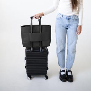 May include: A black rolling suitcase with a matching black tote bag attached. The tote bag has a handle and a strap to secure it to the suitcase. The suitcase has four wheels and a retractable handle. The items are on a white background.