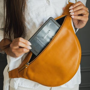 May include: A tan leather fanny pack with a gold zipper and hardware. The bag is open, revealing a Kindle e-reader with the text "Your next great read". The e-reader displays a woman standing near the moon.