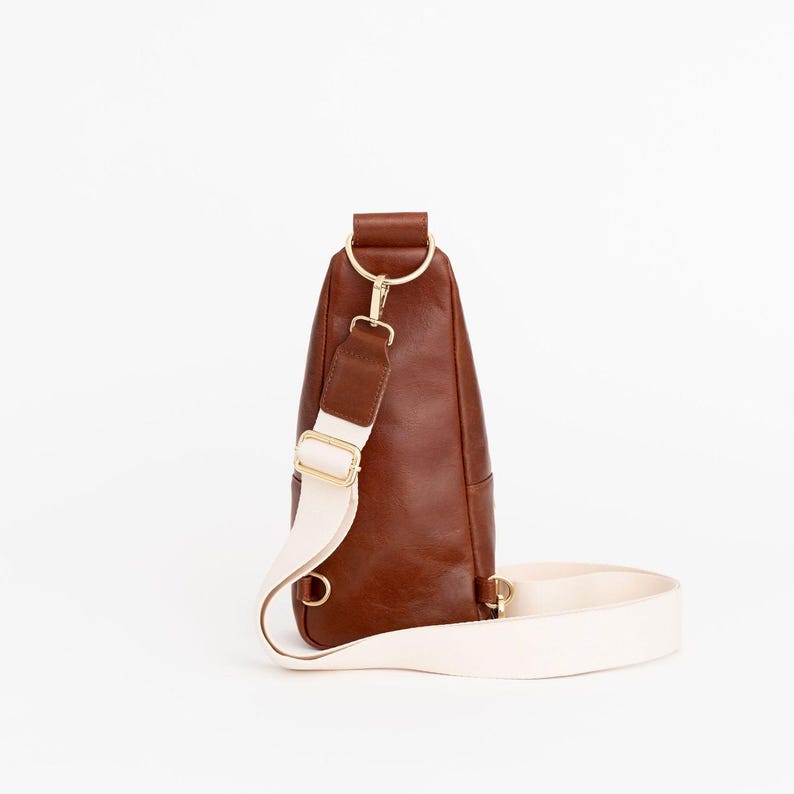 May include: A brown leather sling bag with a cream-colored strap and gold-colored hardware. The bag has a rounded shape and a single strap that can be worn across the body. The bag is set against a white background.