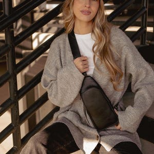 May include: A black leather crossbody bag with a zipper closure and adjustable strap. The bag is worn over a white t-shirt and a gray cardigan. A black baseball cap with the word "GIANTS" in red lettering is worn.