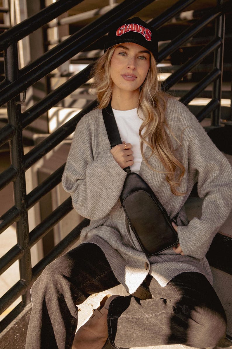 May include: A black leather crossbody bag with a zipper closure and adjustable strap. The bag is worn over a white t-shirt and a gray cardigan. A black baseball cap with the word "GIANTS" in red lettering is worn.